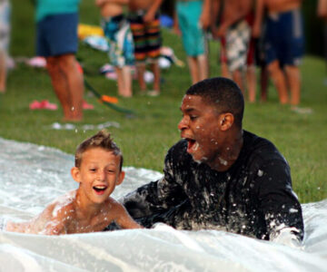 A junior boy camper speeds down our slip n' slide with his cabin counselor.
