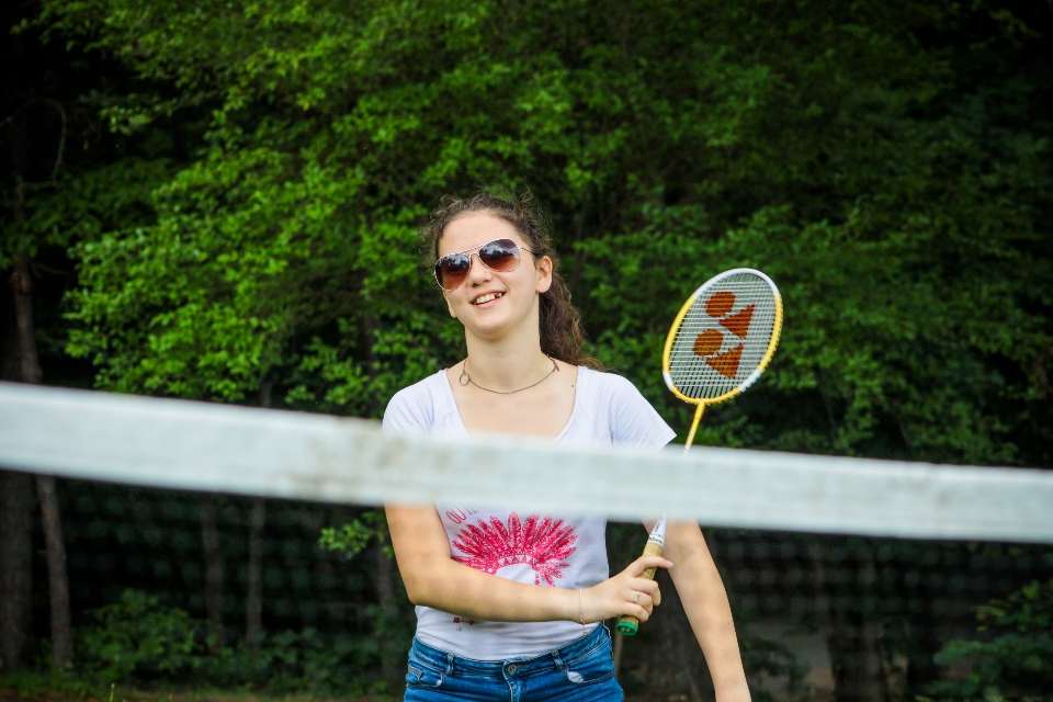 A teen camper smiles holding a badminton racket behind the net during our residential camp.