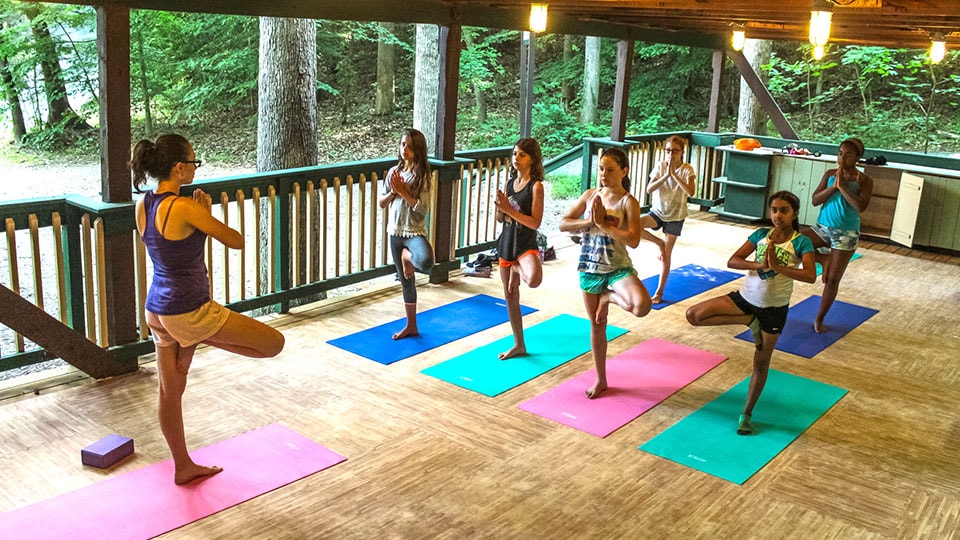 A counselor leads a group of junior and teen campers in a yoga stance on colored mats in our Camp Friendship dance pavilion.