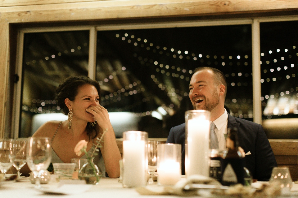 A tablescape of candles, lights, and flowers fill this shot of a summer camp wedding, as the bride and groom smile and laugh in the background.