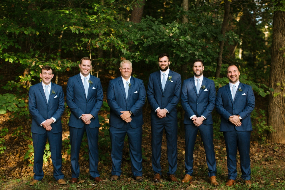 A group of groomsmen and the groom, all dressed in navy suits, pose in the natural tree line at our Virginia wedding venue.