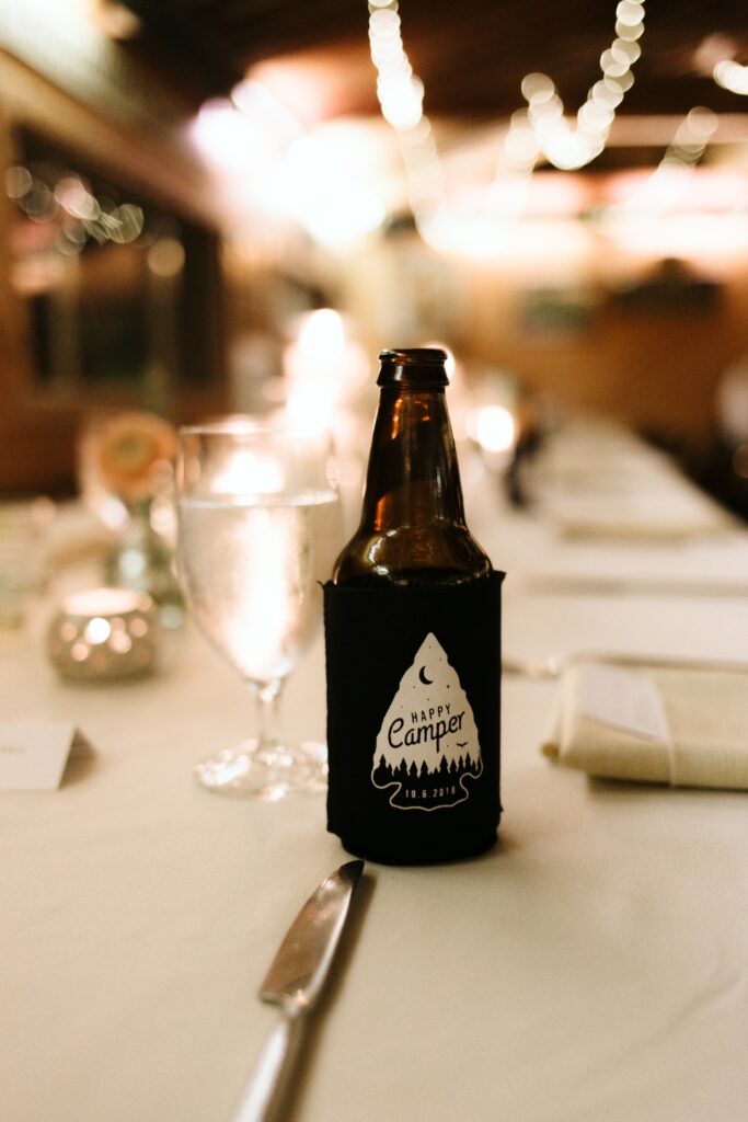 A bottle of beer with a camp beer cozy sits on a decorated camp table set with a white table cloth for the wedding.