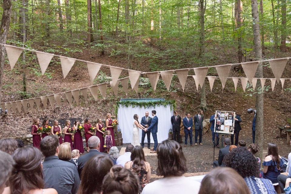 Summer Camp wedding with bride, groom, bridesmaids, groomsmen, and crowd watching the ceremony with bunting hanging above.