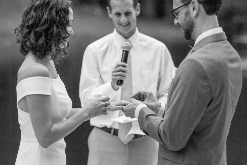 Groom places wedding band on bride's finger with officiate looking on.