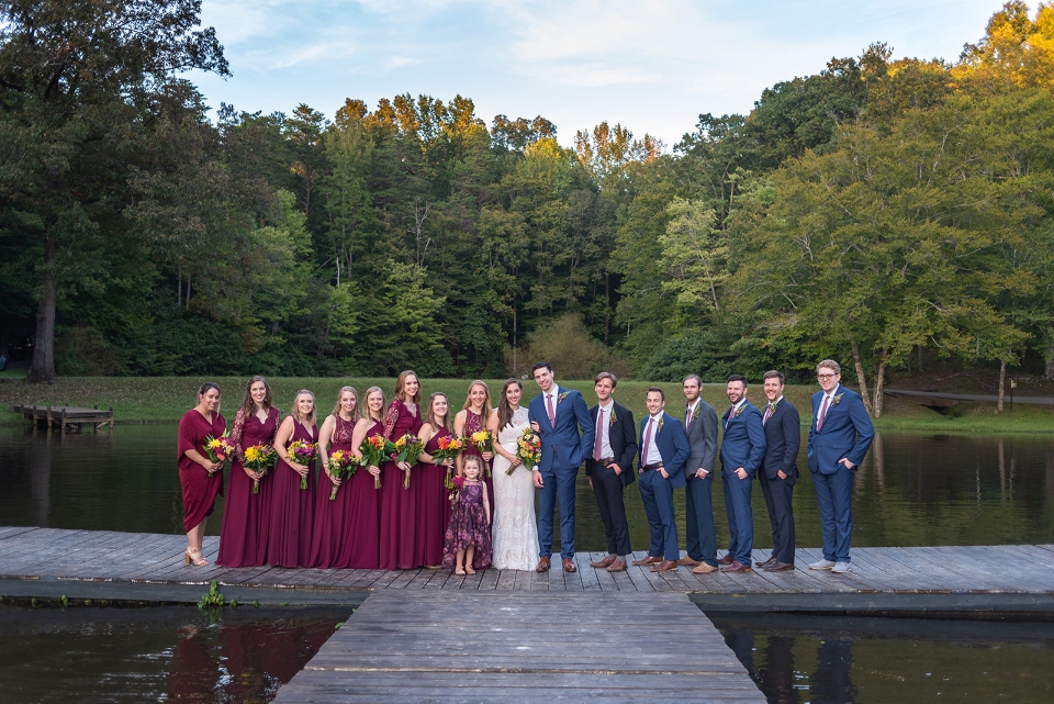 A wedding party of brides maids and groomsmen stand on the dock at the lake with the forest rising behind them at our friendship lake.
