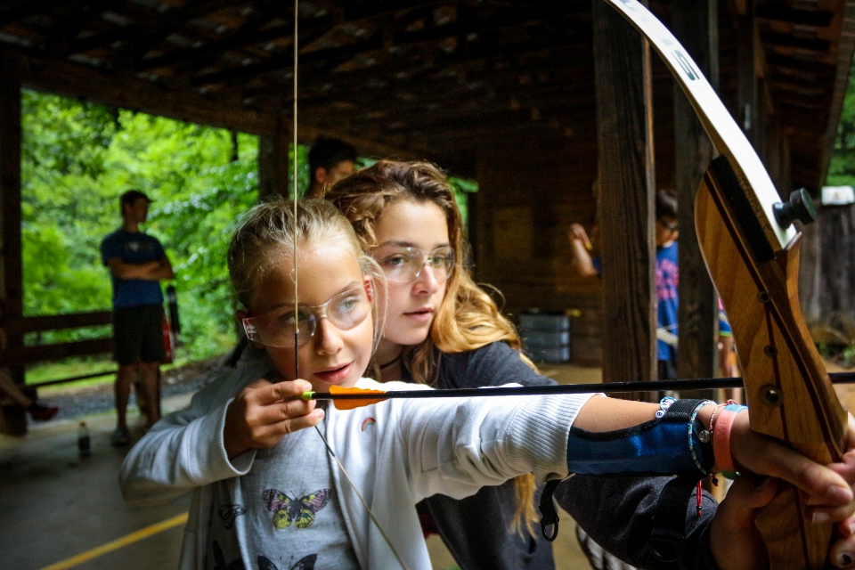 A counselor helps a junior camper aim her first shot on the archery range.