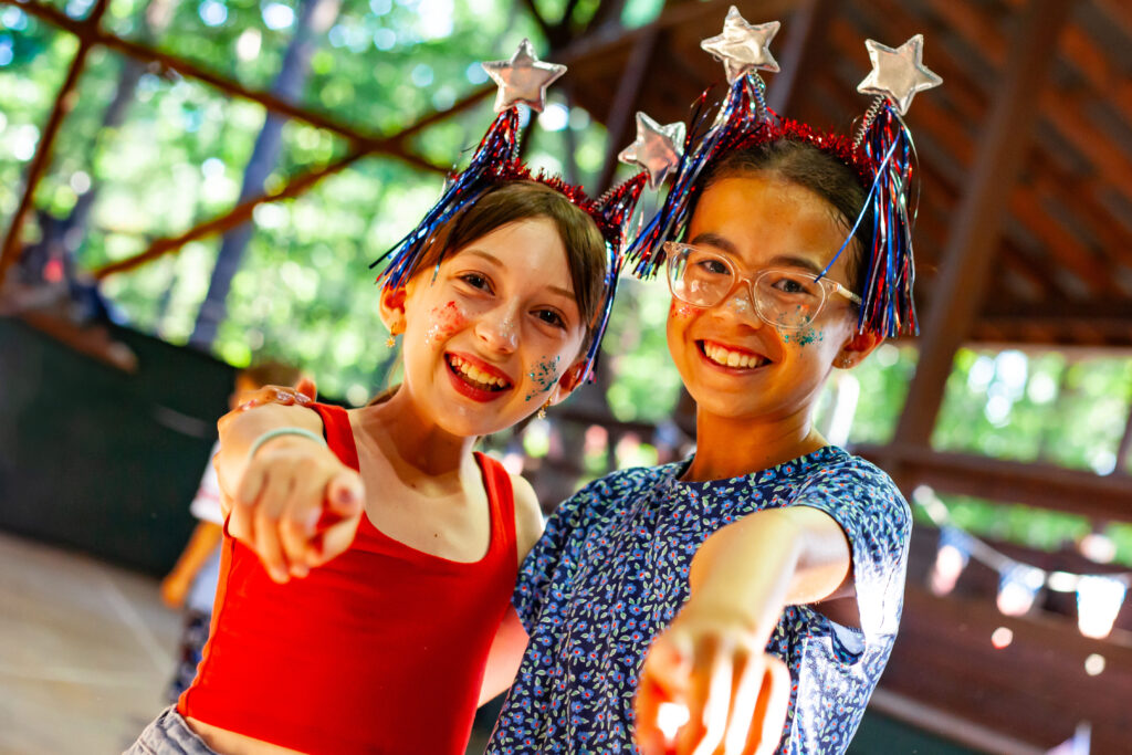 2 girls in red, white, and blue shirts and headbands pointing at the camera and smiling
