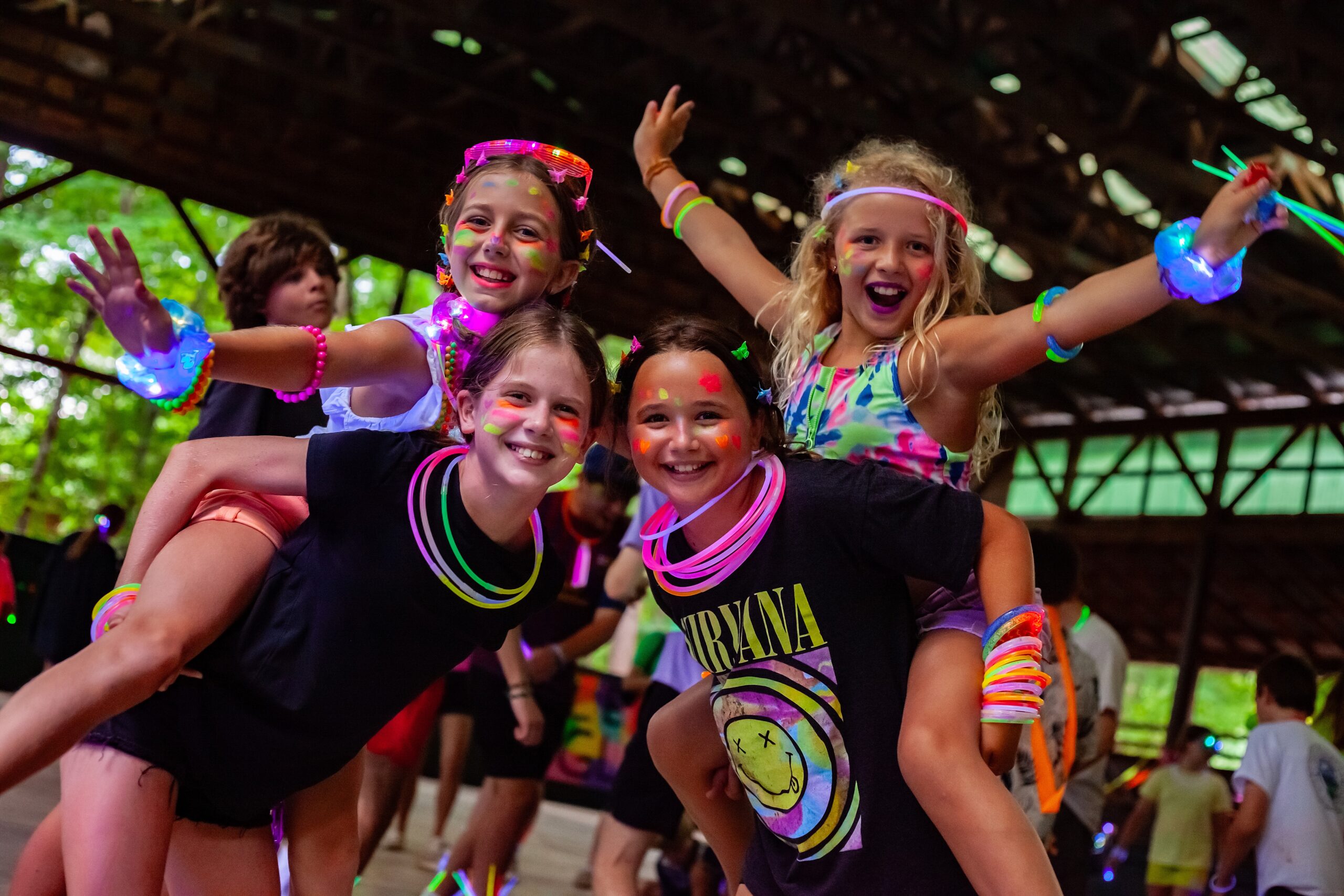 4 girls with neon clothing and bracelets on smiling at the camera. 2 of the girls are giving the other 2 piggyback rides