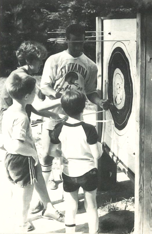 A black-and-white photo of a man showing children how to pull arrows from a target